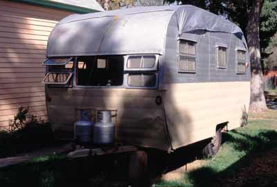 Image of old teardrop trailer with a tarp covering the top next to a house in a 
residential neighbohood.